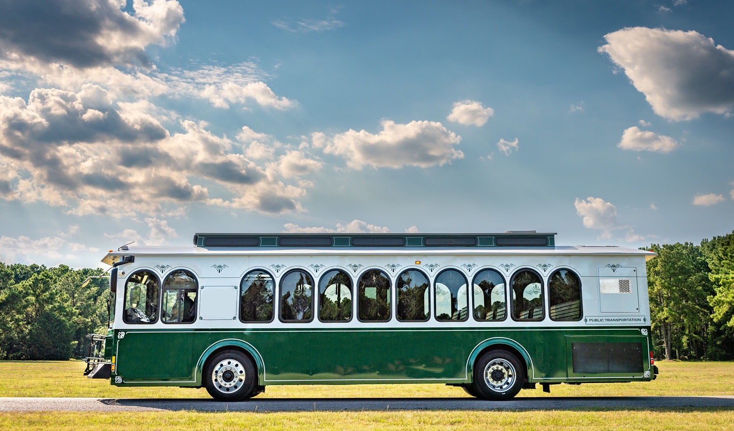  A green and white trolley bus with arched windows and decorative trim is parked on a paved road surrounded by lush greenery under a partly cloudy sky. The trolley features a classic design with a modern touch, combining vintage charm with contemporary public transportation features. The sunlight highlights the vehicle's polished exterior, emphasizing its elegant craftsmanship.