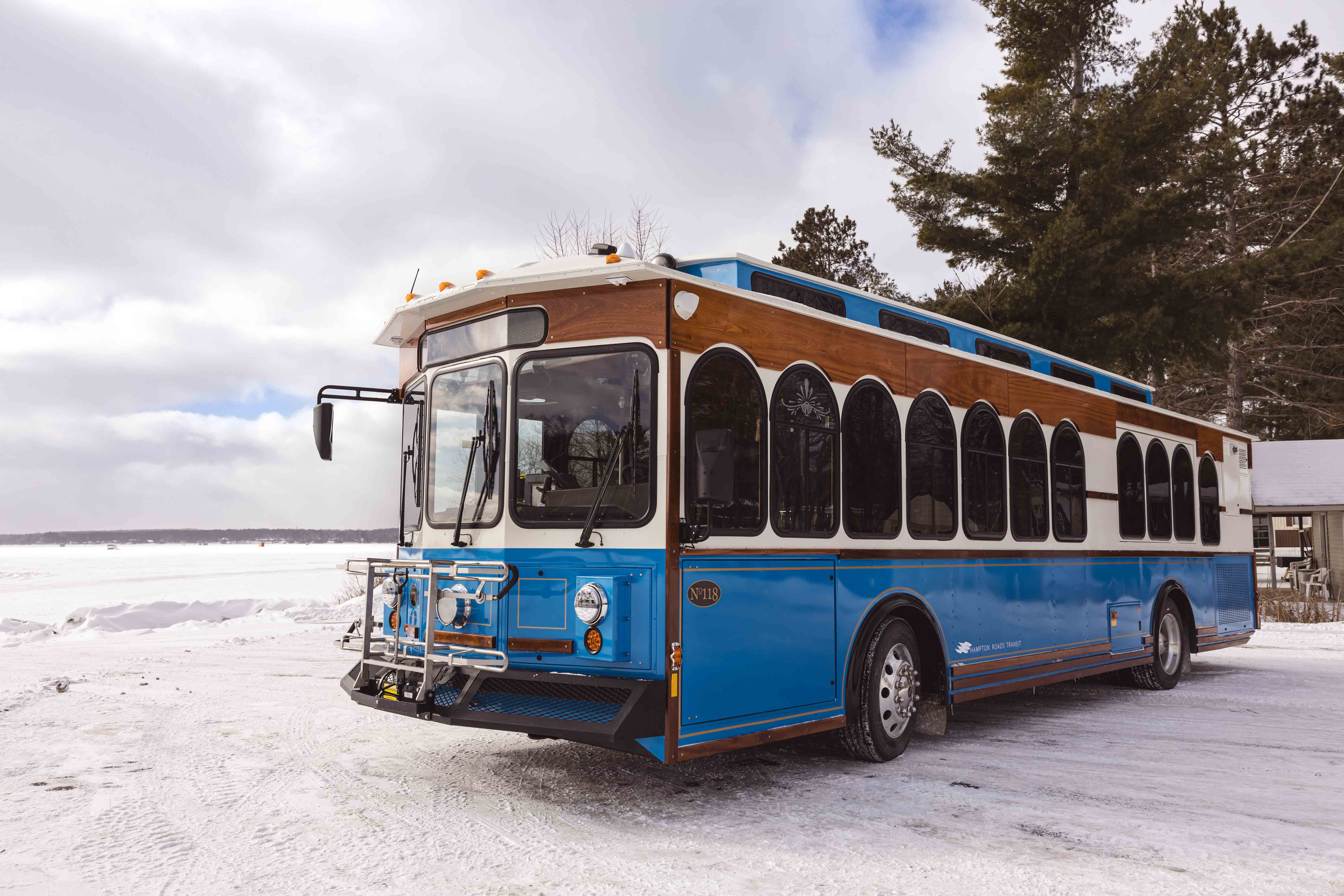 Text: A blue and white trolley with wood panel accents is parked on a snowy landscape with a frozen lake and evergreen trees in the background. The trolley features arched windows, a front bike rack, and a classic design that blends vintage charm with modern transit functionality. The winter setting highlights the trolley’s durability and year-round usability.