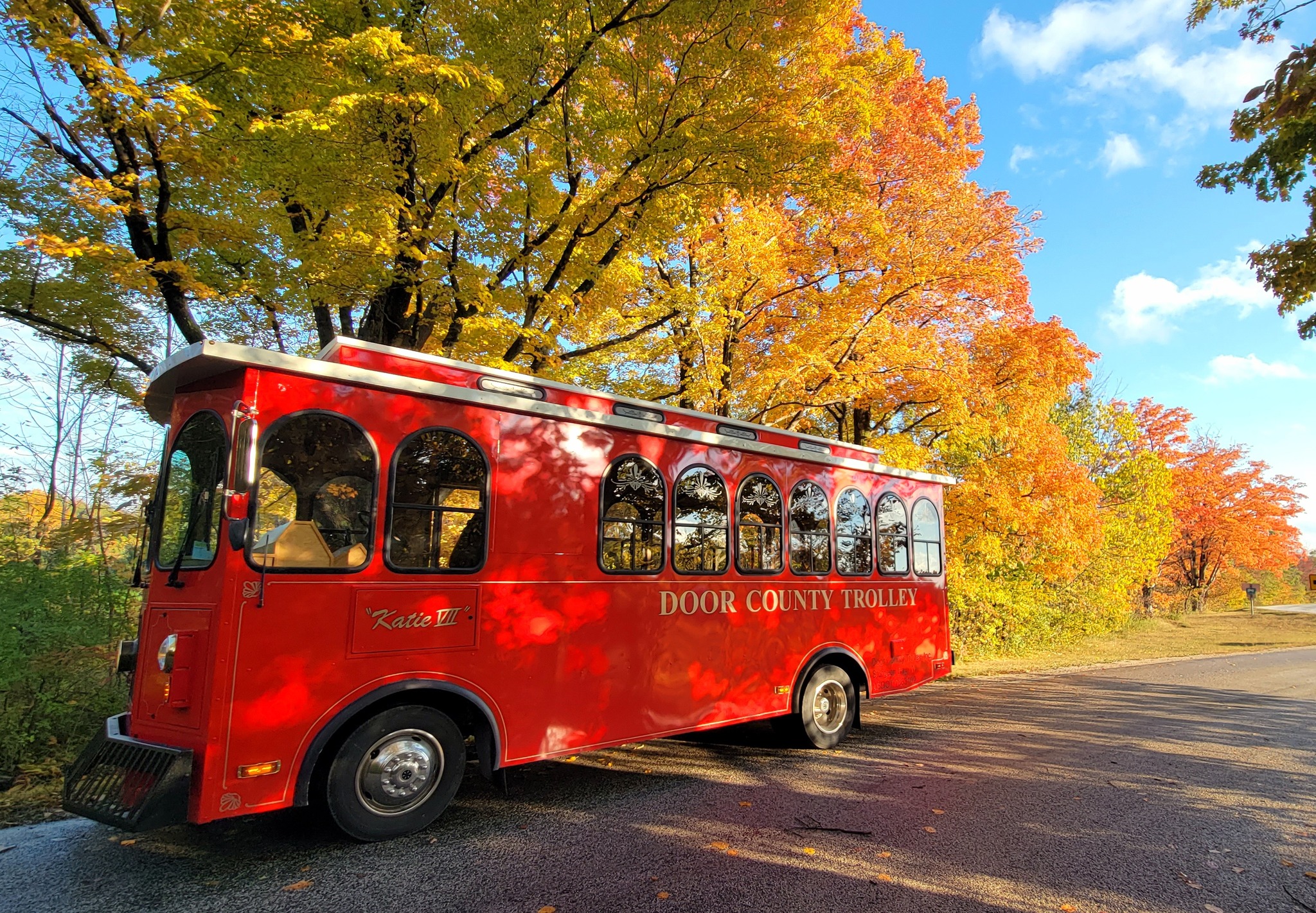 Bright red Door County Trolley parked on a quiet road surrounded by vivid fall foliage, with golden and orange leaves under a clear blue sky.