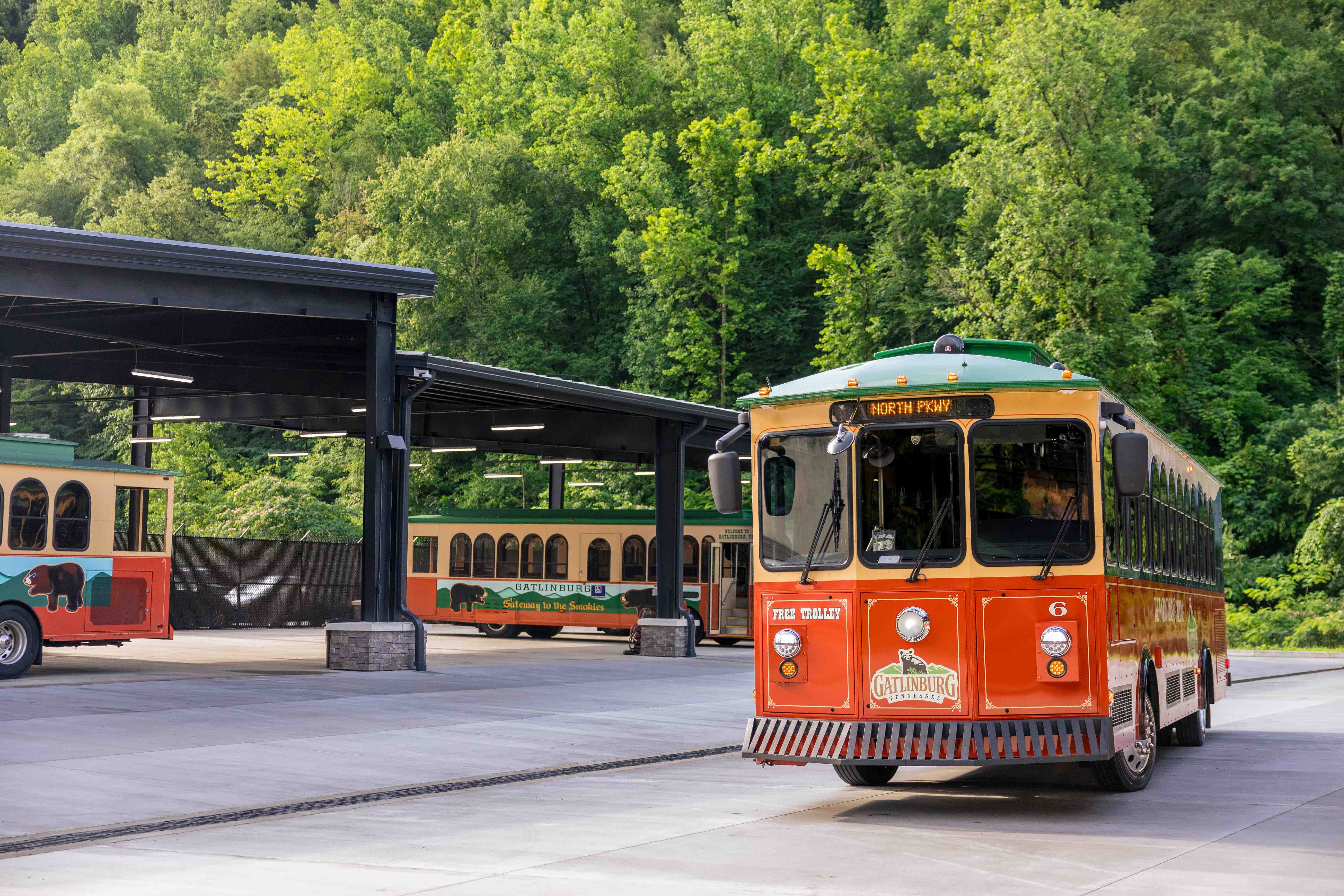 Three Gatlinburg trolleys parked at a transit facility surrounded by lush green trees. The orange and green trolley in the foreground displays "North Pkwy" on its sign and "Free Trolley" above the bumper. Two additional trolleys are visible under the shelter in the background.