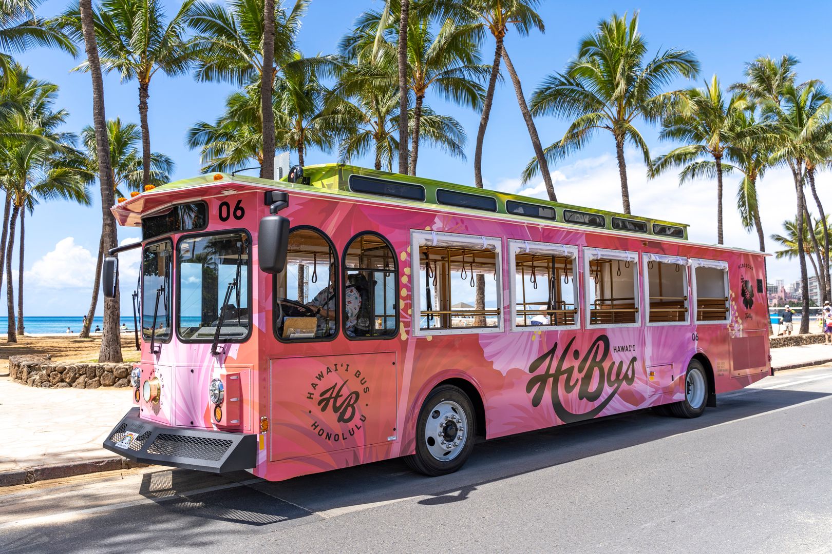 right pink Honolulu “HiBus” trolley parked along a palm tree-lined street near the beach, showcasing its custom exterior design.