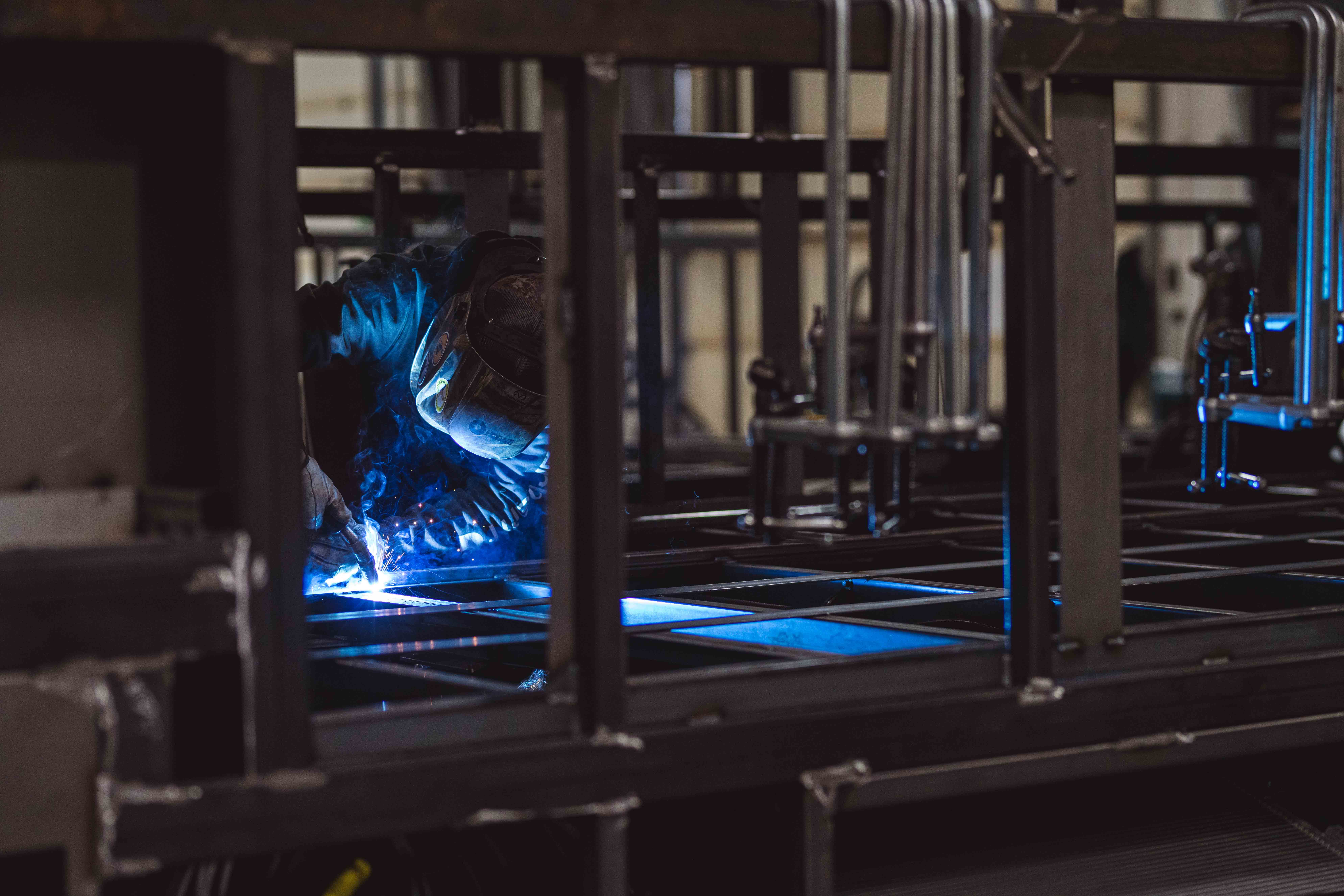 Welder working on a steel frame inside a manufacturing facility, sparks and blue light reflecting off the metal structure during the fabrication process.