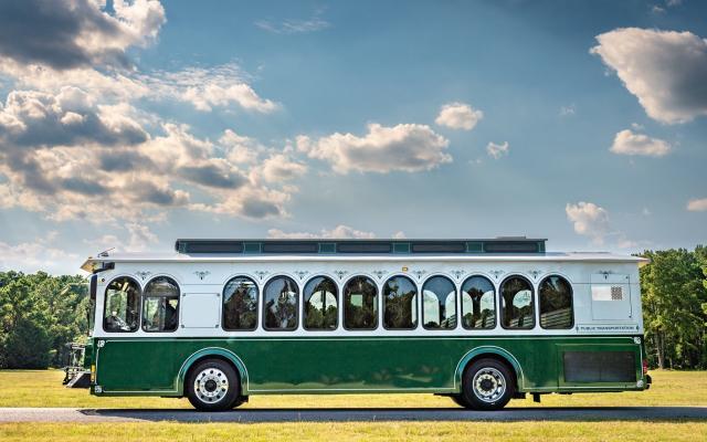  A green and white trolley bus with arched windows and decorative trim is parked on a paved road surrounded by lush greenery under a partly cloudy sky. The trolley features a classic design with a modern touch, combining vintage charm with contemporary public transportation features. The sunlight highlights the vehicle's polished exterior, emphasizing its elegant craftsmanship.