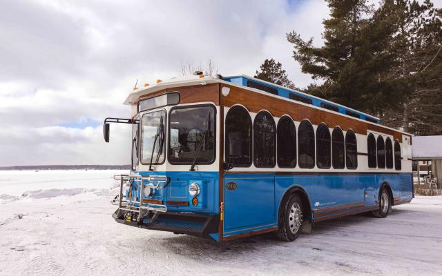 Text: A blue and white trolley with wood panel accents is parked on a snowy landscape with a frozen lake and evergreen trees in the background. The trolley features arched windows, a front bike rack, and a classic design that blends vintage charm with modern transit functionality. The winter setting highlights the trolley’s durability and year-round usability.