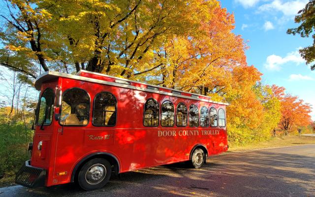  Bright red Door County Trolley parked on a quiet road surrounded by vivid fall foliage, with golden and orange leaves under a clear blue sky.