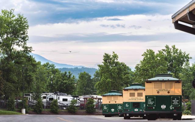 Three green and cream Pigeon Forge trolleys parked in a lot with the Smoky Mountains and a cloudy sky in the background.
