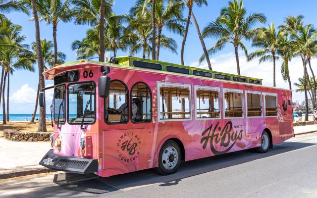 right pink Honolulu “HiBus” trolley parked along a palm tree-lined street near the beach, showcasing its custom exterior design.