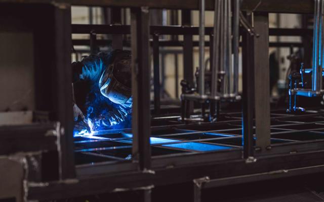 Welder working on a steel frame inside a manufacturing facility, sparks and blue light reflecting off the metal structure during the fabrication process.